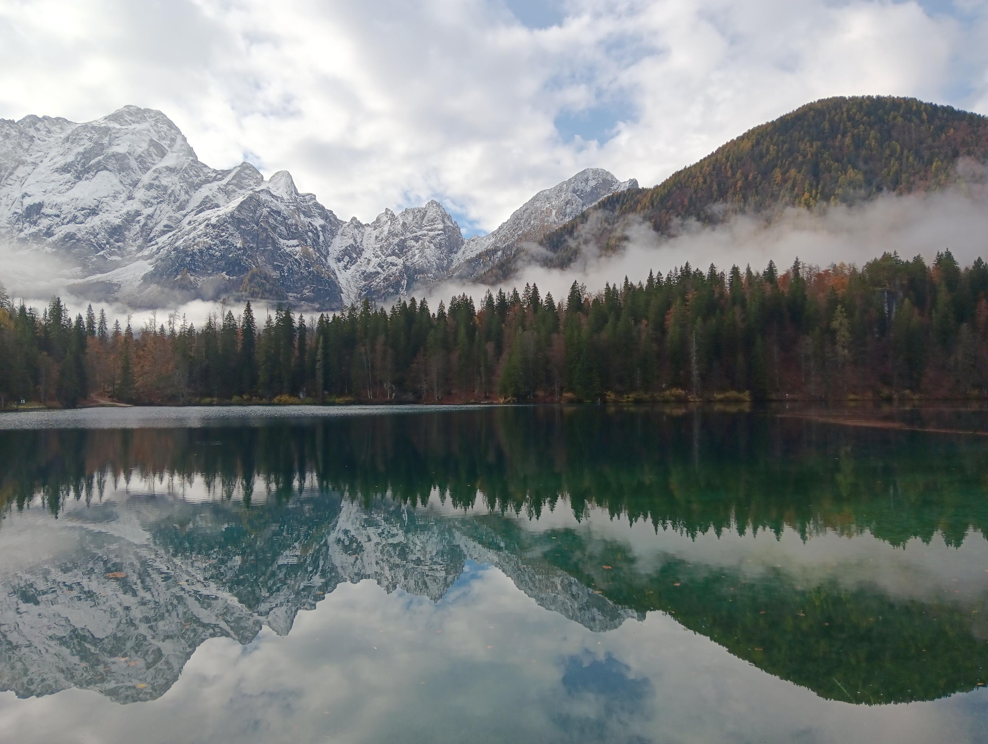 Mountain tops slightly covered with snow on the left, a hill with mixed trees with red, green, yellow and orange colored leaves on the right. Before that is a thick fog trail and more trees. Everything is reflected in a lake with clear water. On the right, the water is more shallow, resulting in a very slight greenish tint for that half of the reflection.