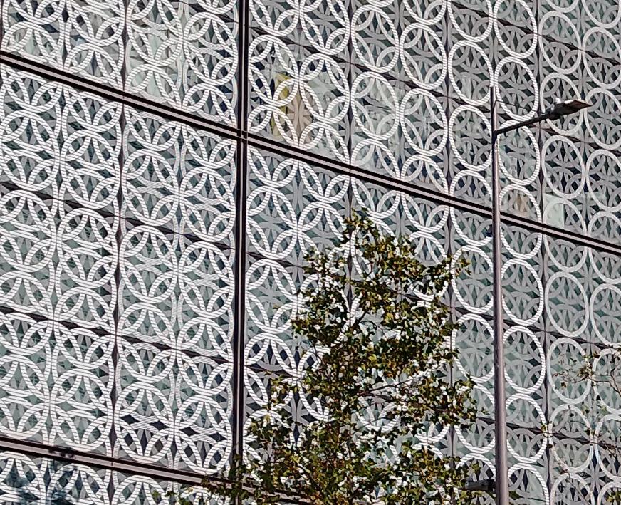 The faced of a building is dominated by white overlapping circles in two layers. Part of a tree and lamp post are visible. A bird sits on top of the lamp post.
The front layer circles have thin horizontal stripes. From left to right, the front layer has fewer circles and reveals more of the background layer of circles.