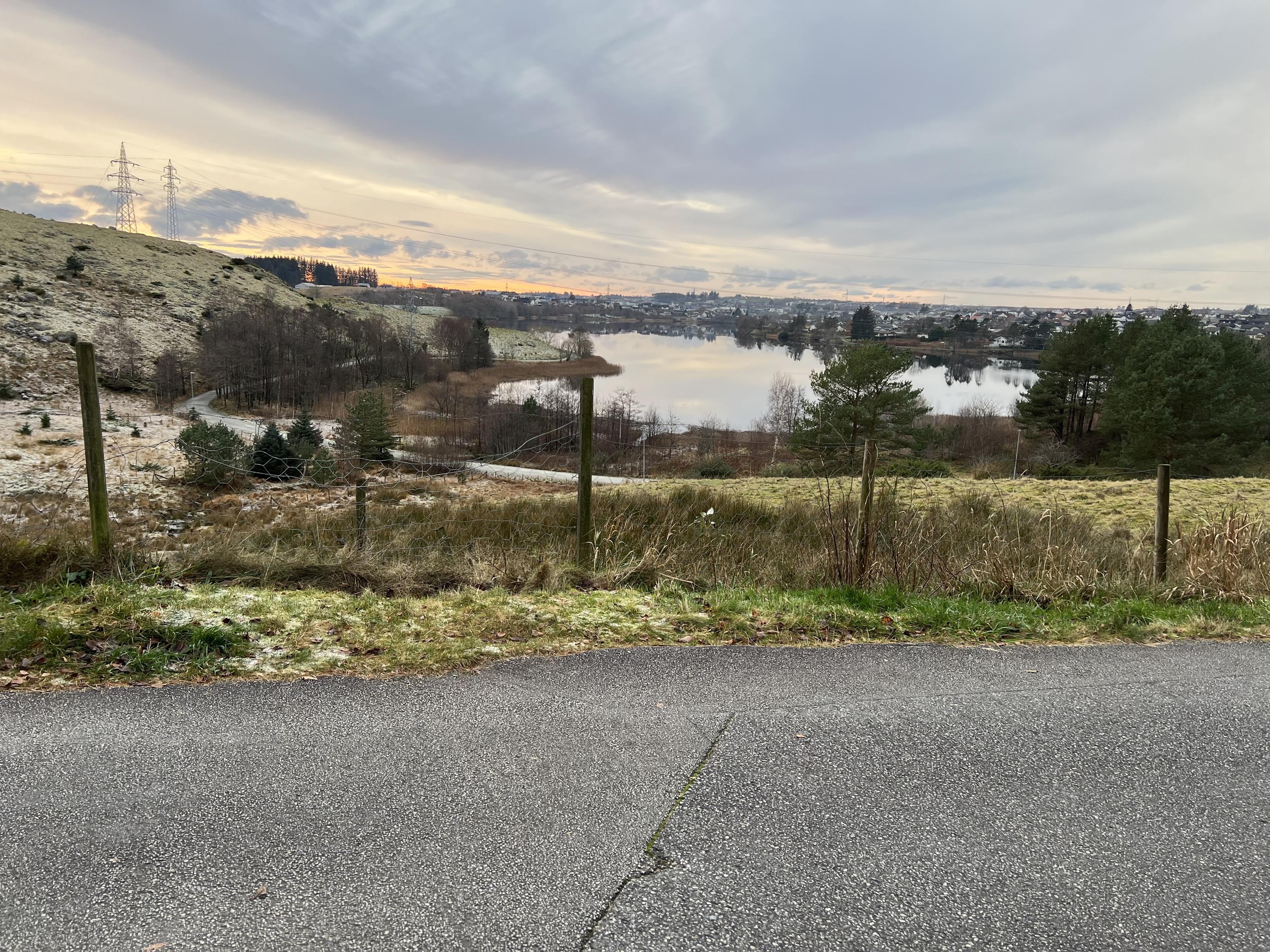 A scenic landscape featuring a lake surrounded by trees and hills, with a gravel road leading to the water. The sky displays soft colors of sunset, and power lines are visible in the background. A fence marks the foreground, with patches of grass and frost