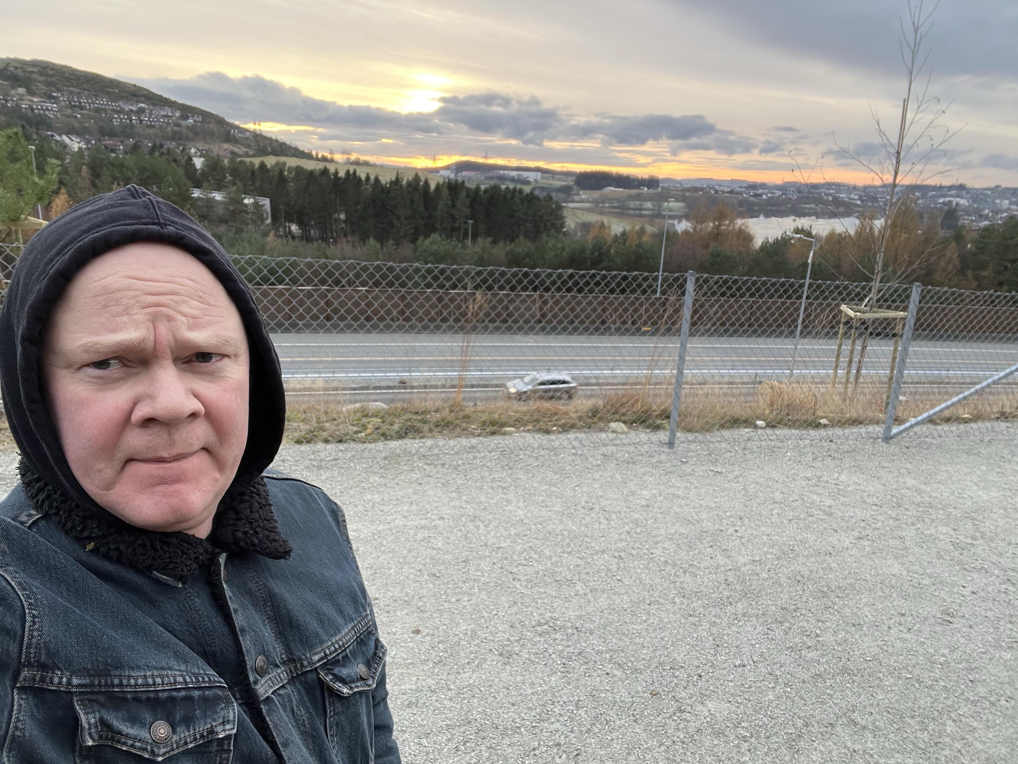 A person stands in the foreground, wearing a hooded jacket and a denim jacket. In the background, a landscape features trees, a winding road, and a sunset with clouds and a hills. A fence is present near the road.