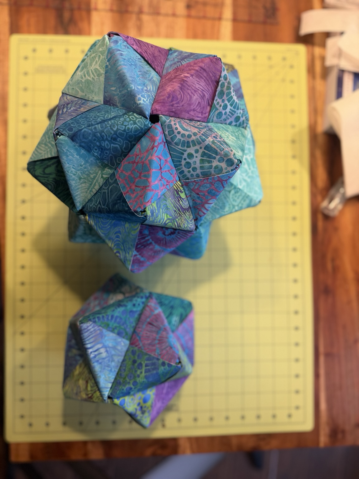 Origami fabric icosahedronand octahedron side-by side with turquoise purple and blue fabrics on a cutting mat