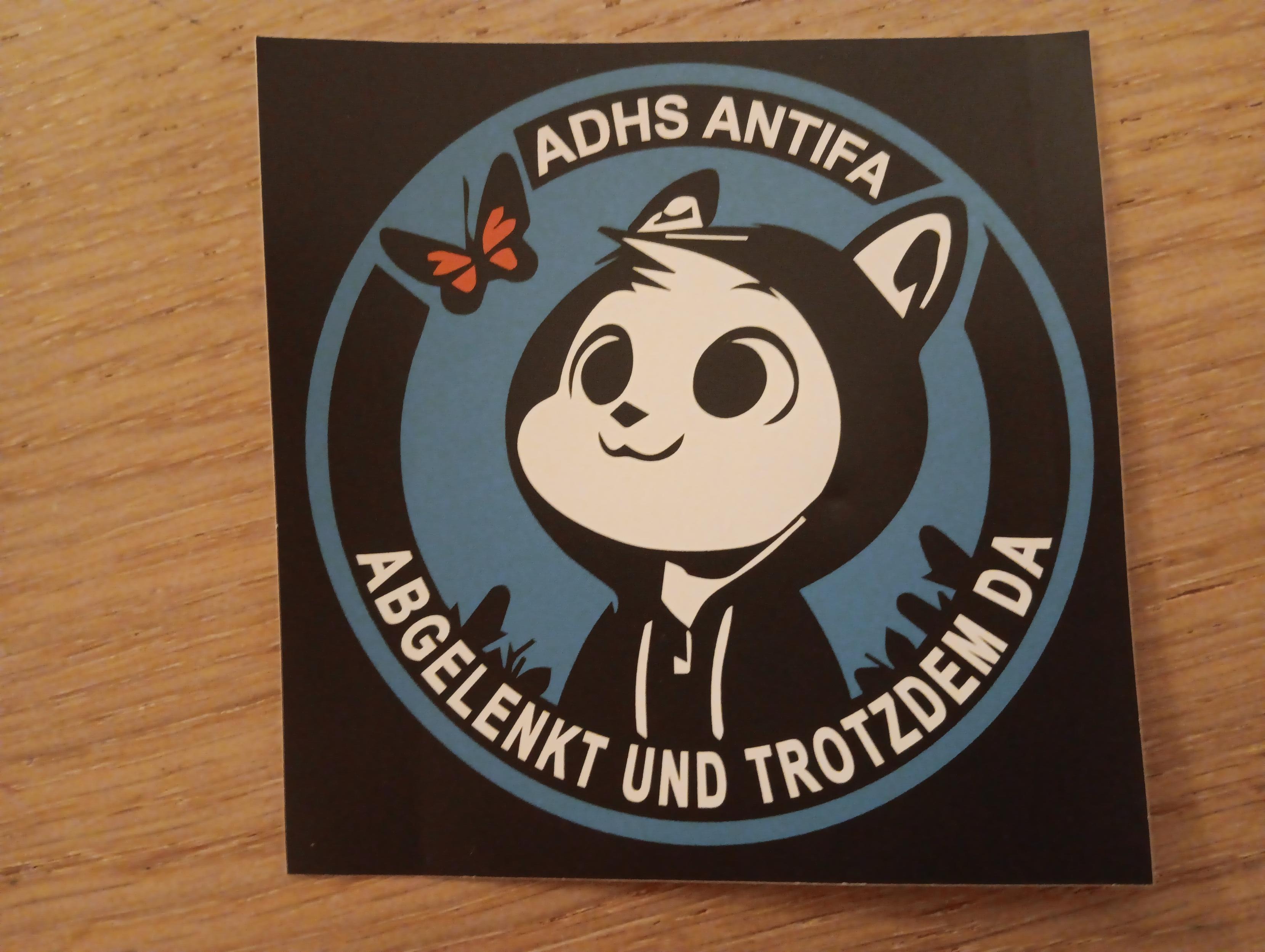 Quadratischer Sticker mit rundem Motiv, mit der Aufschrift "ADHS Antifa" oben im Kreis, "abgelenkt und trotzdem da" unten im Kreis, in Groรbuchstaben. In der Mitte ist die obere Hรคlfte einer, wie ein Mensch stehende, weiรes Katze in schwarzem Hoodie, die abgelenkt auf einen orange-schwarzen Schmetterling schaut. Der Sticker liegt auf einem braunen Spanplatten-Tisch mit klar erkennbarer Maserung.