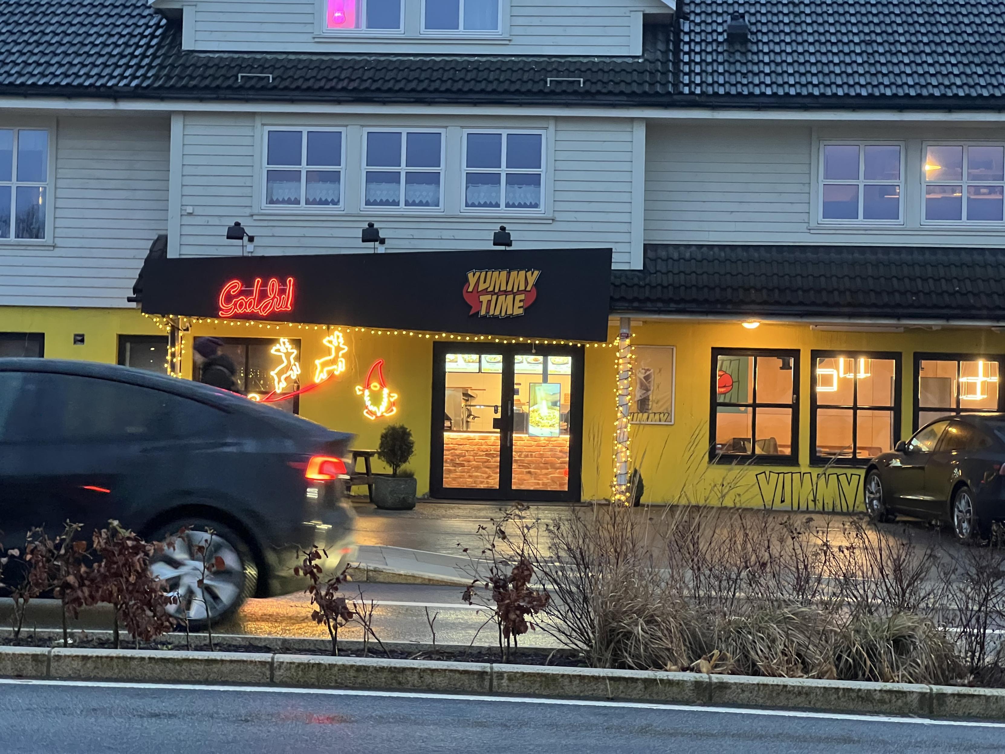 The image shows a building with two distinct storefronts. One side features a neon sign reading "God Girl" with holiday decorations, while the other side displays "Yummy Time" in bold lettering. There are large windows and a few cars parked outside