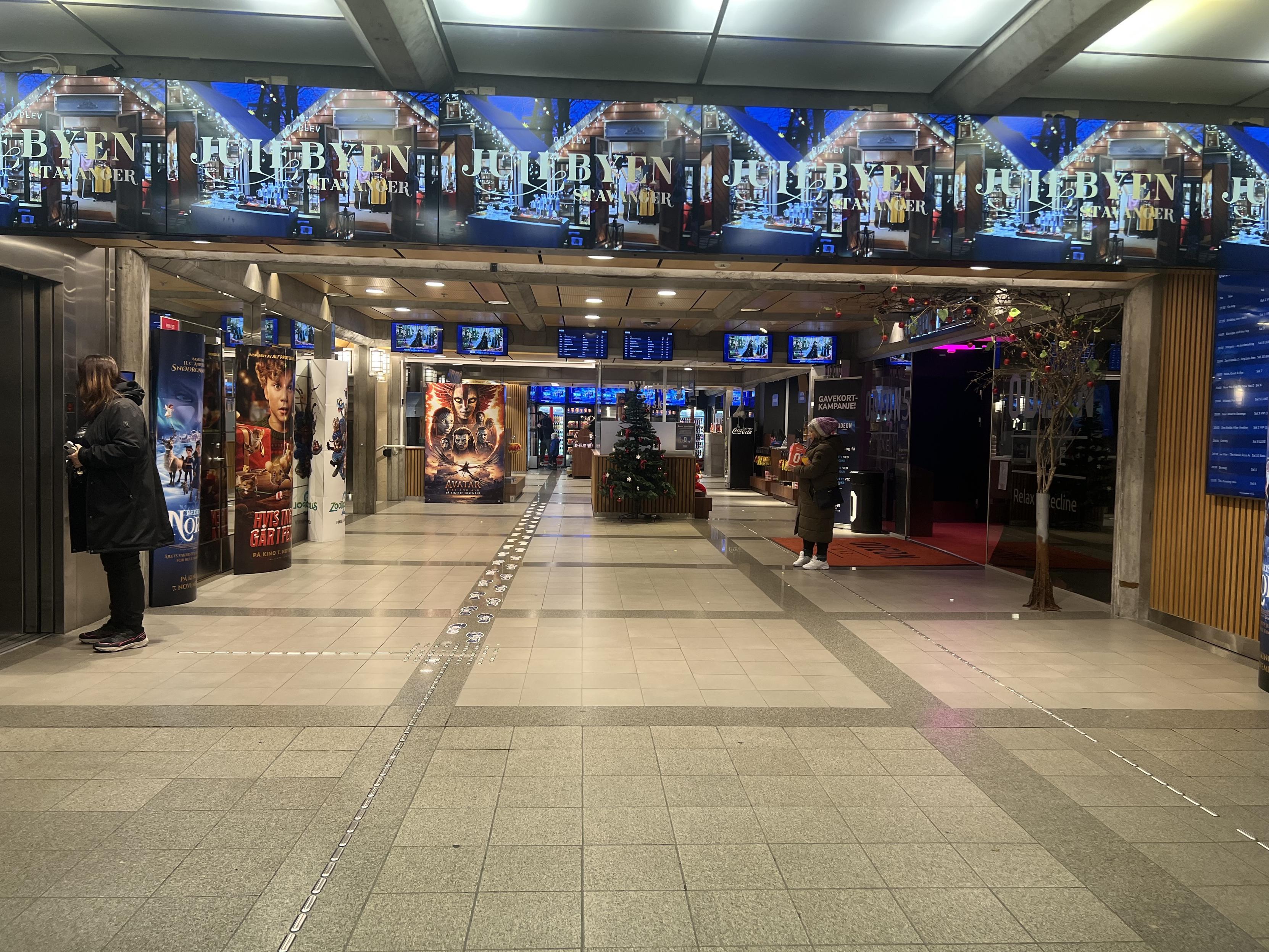 The image shows a modern indoor space, likely a cinema or entertainment venue, featuring various movie posters along the walls. There's a seating area, digital screens displaying schedules, and a Christmas tree. People are seen waiting and interacting in the space. The overall