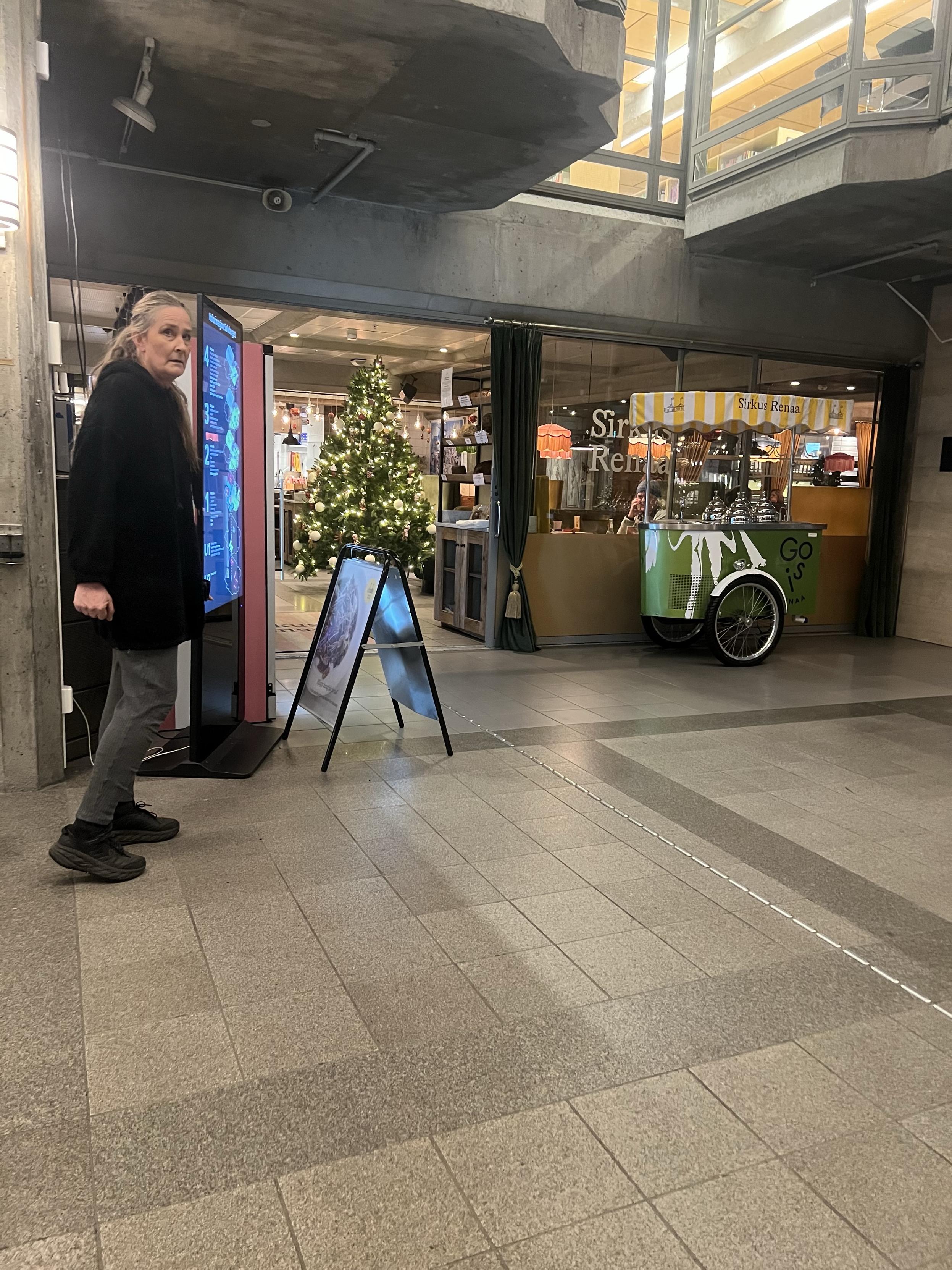 An indoor space featuring a woman in a black coat standing near a digital display. There's a decorated Christmas tree in the background, and a food cart nearby with a striped awning. The space has modern architectural elements and visible retail areas.