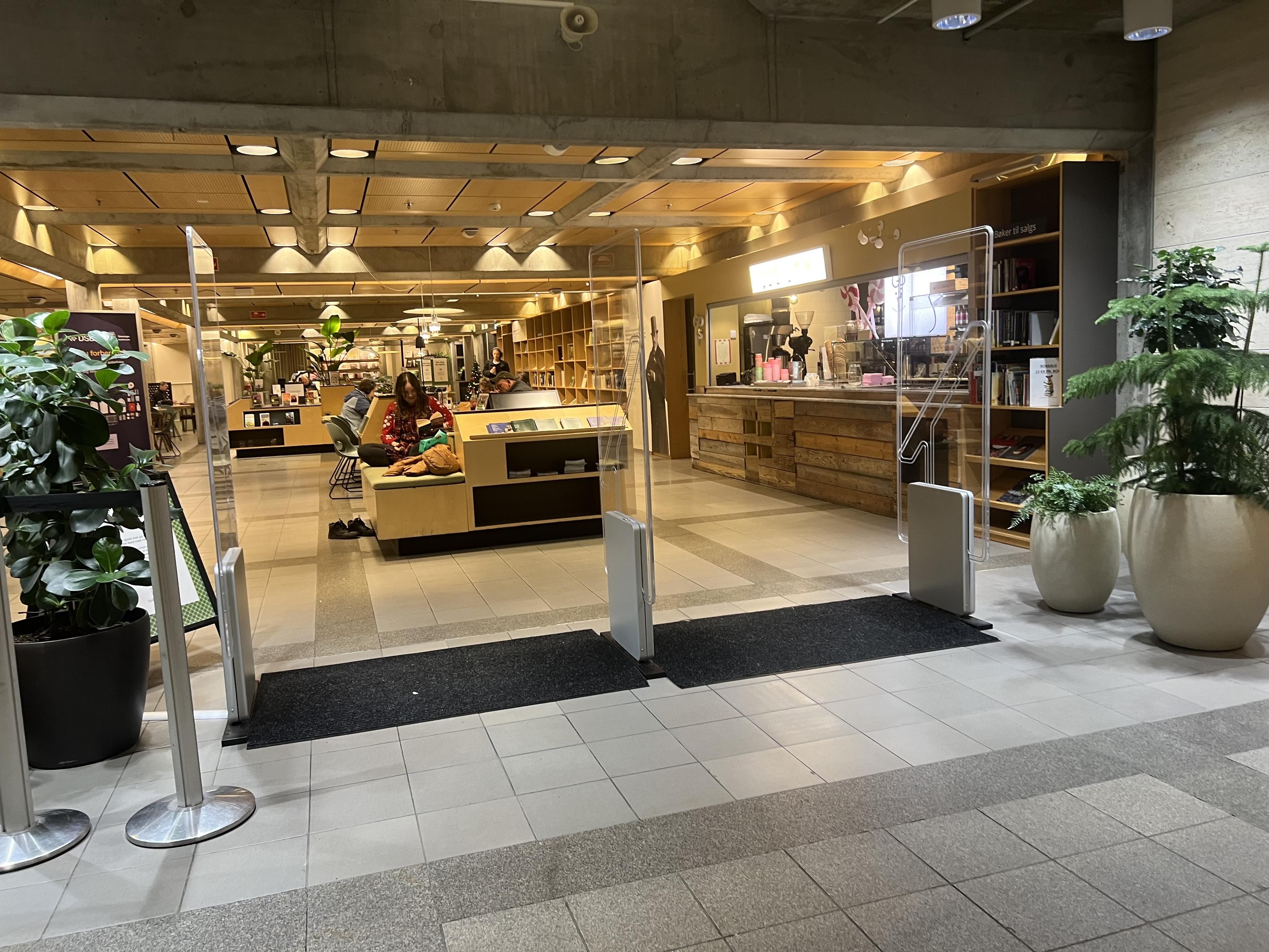 A modern indoor space featuring a reception area with wooden fixtures, a café counter, and a seating area. There are plants and bookshelves visible, with a few people sitting and using devices. The entrance is marked by transparent barriers.