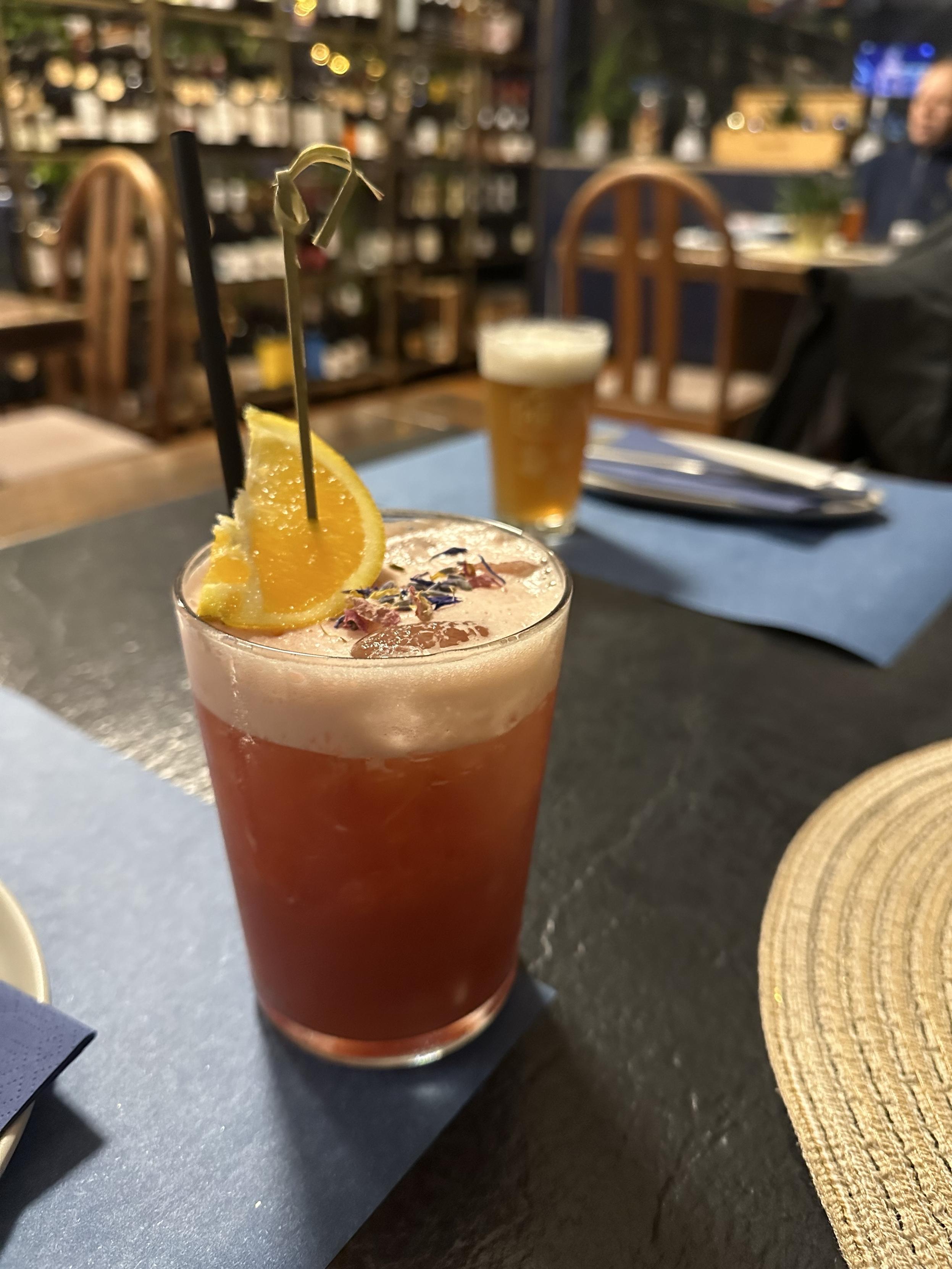 Fancy-looking pink mocktail with foam, decorated with an orange wedge and dried rose petals and lavender. In the background a beer, chairs, tables and a wine rack full of wine bottles.