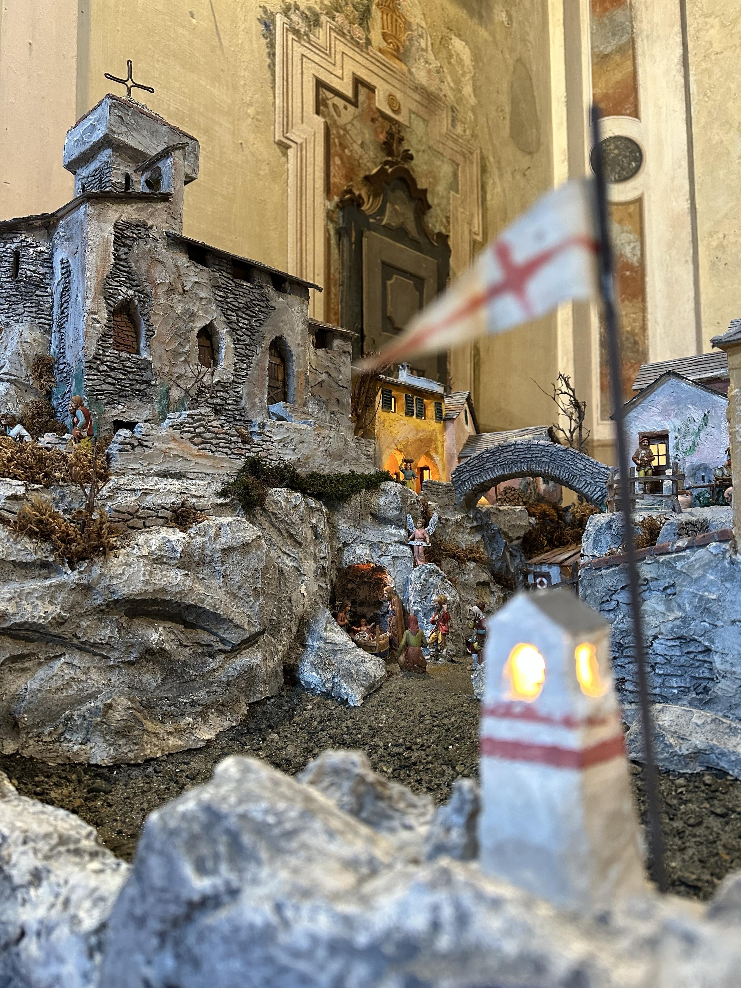 A miniature town housings nativity scene, built inside a church. In the foreground out of focus a tiny white lighthouse with a flag on top.