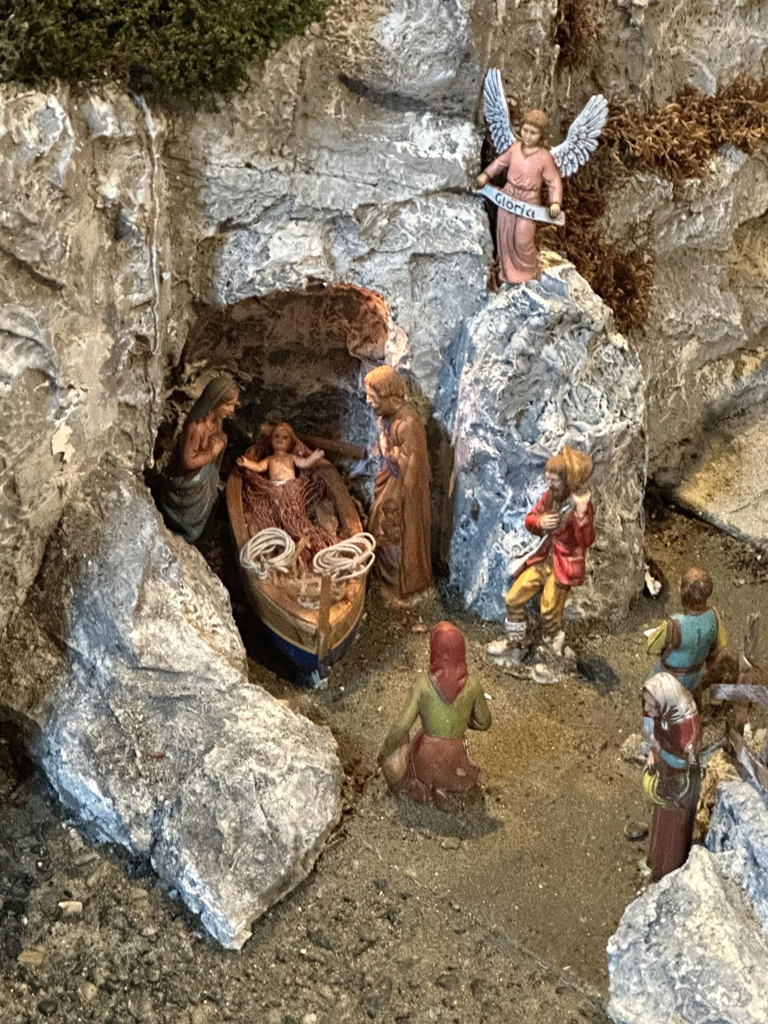 A miniature nativity scene with small figurines. Baby JC is in a boat inside an opening in the rock face, with Mary and Joseph looking over him. On top an angel with a banner that says “Gloria”. Some worshippers have gathered.
