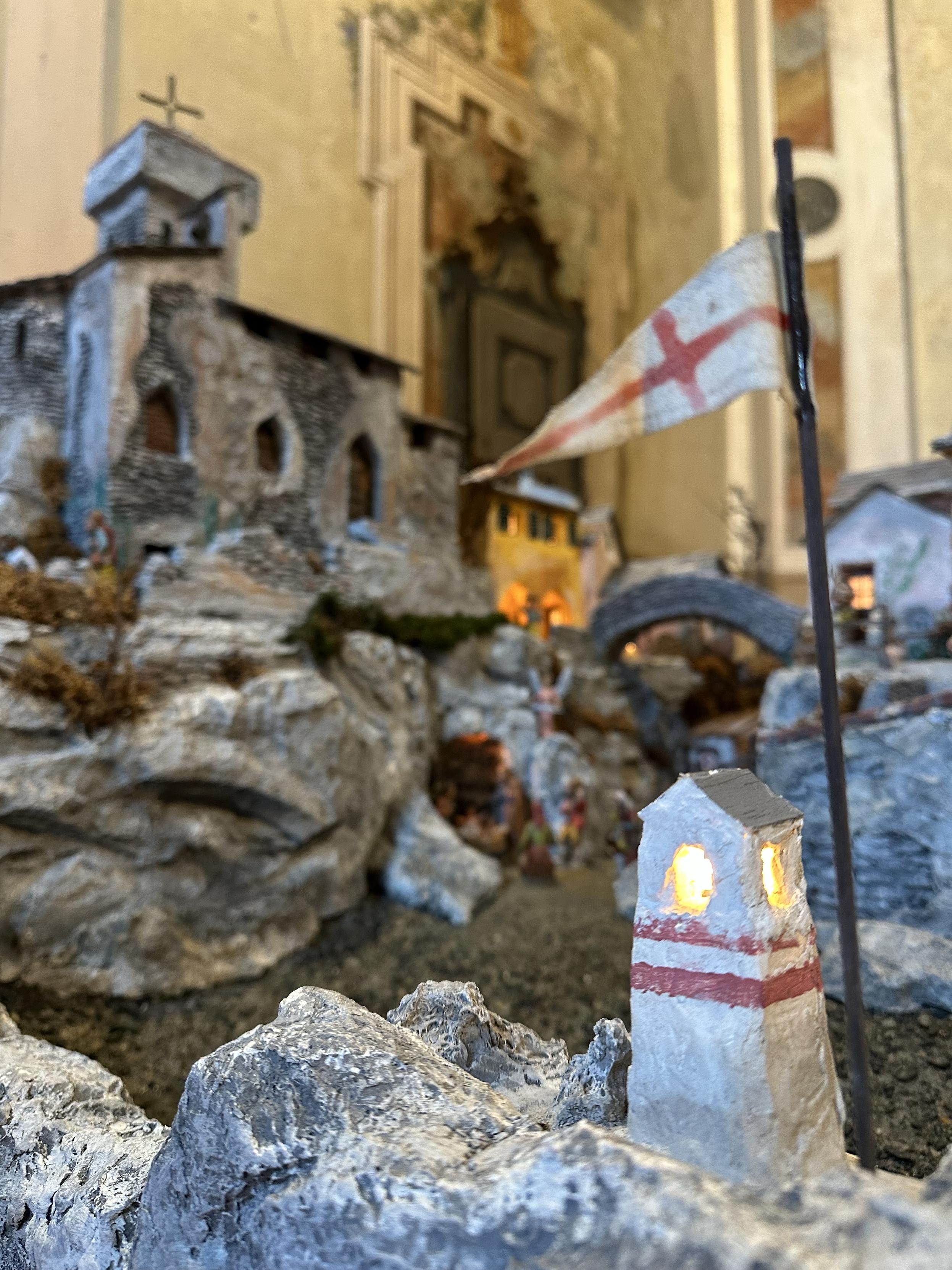 Same view of the miniature village built inside the church, but now it is out of focus and the white lighthouse is in focus. It has two horizontal red stripes right below its light and the white triangular flag on top has a Red Cross.