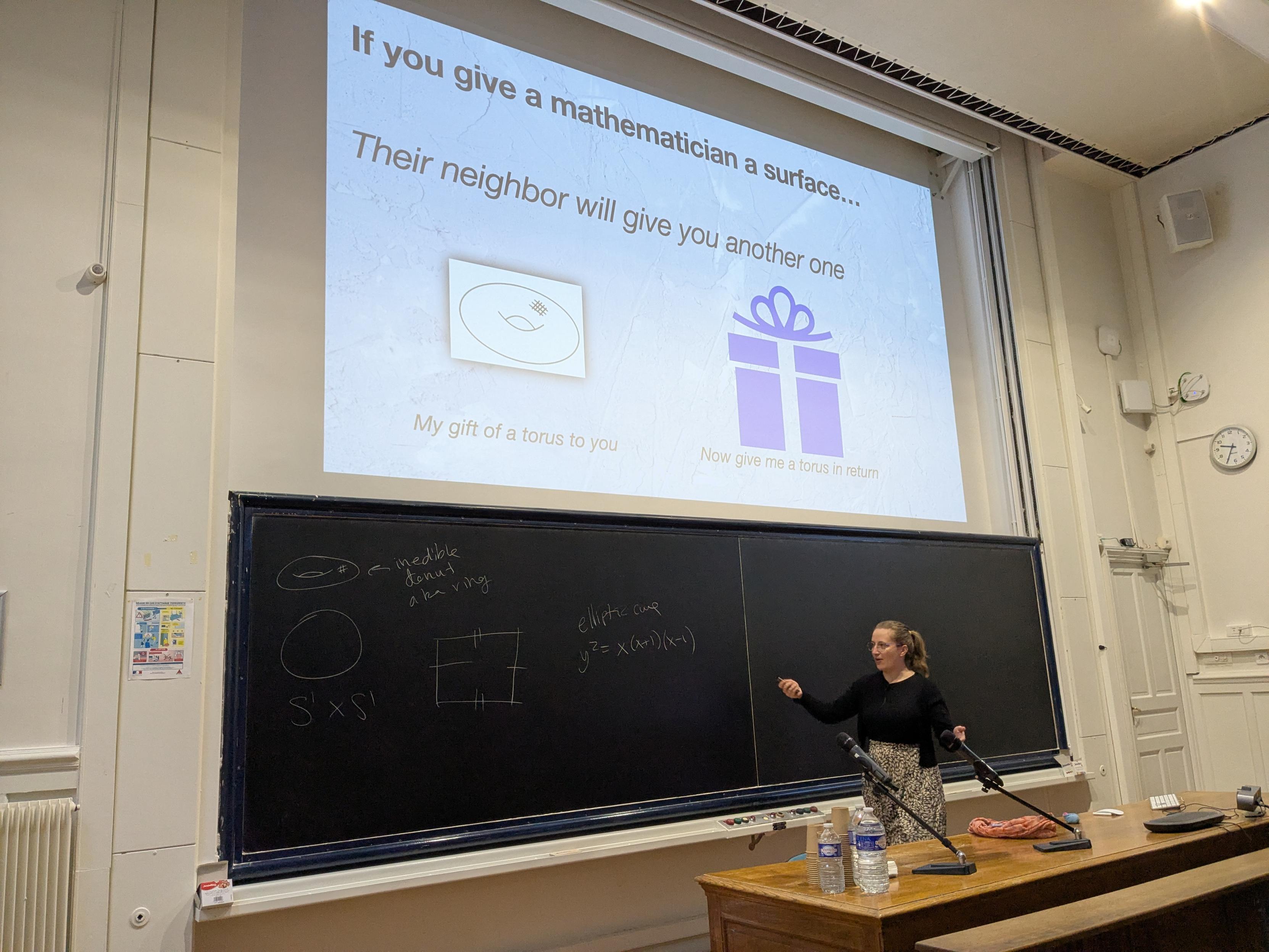 A person, Samantha Fairchild, stands in a lecture hall pointing toward a large blackboard. They have blonde hair tied back and are wearing a black top with a patterned skirt. The blackboard contains hand-drawn diagrams of a torus and a square, along with the equation for an elliptic curve: $y^2 = x(x+1)(x-1)$. Above them, a projector screen displays a slide titled "If you give a mathematician a surface..." featuring a drawing of a torus and a purple gift box icon.