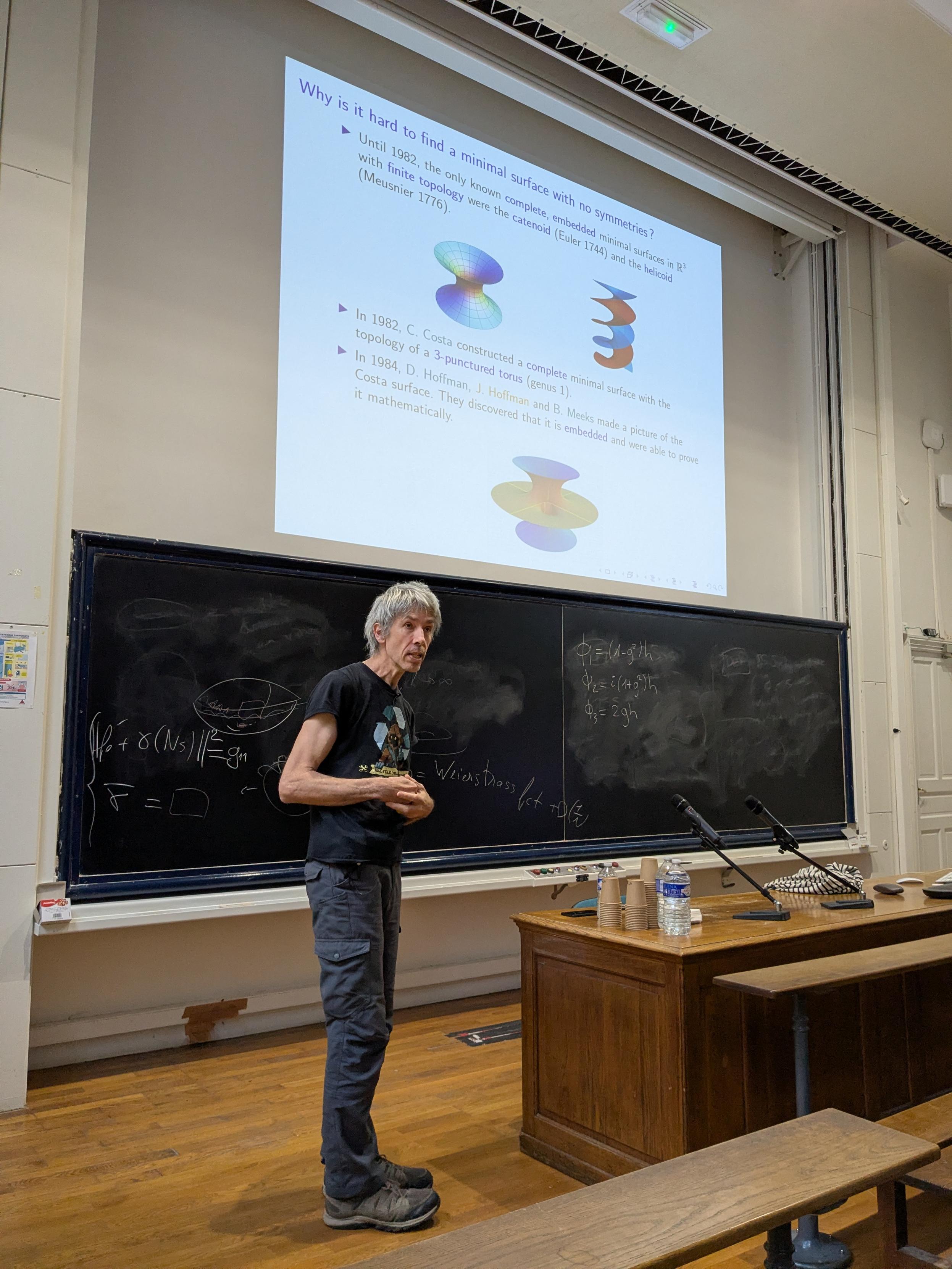A person, Martin Traizet, stands in front of a blackboard covered in mathematical derivations. They have grey hair and are wearing a black graphic t-shirt and grey cargo pants. The blackboard includes formulas for the Weierstrass representation of minimal surfaces, such as $\Phi_1 = (1-g^2)\eta$, $\Phi_2 = i(1+g^2)\eta$, and $\Phi_3 = 2g\eta$. The projector screen above displays a slide titled "Why is it hard to find a minimal surface with no symmetries?" with diagrams of a catenoid, a helicoid, and a Costa surface.