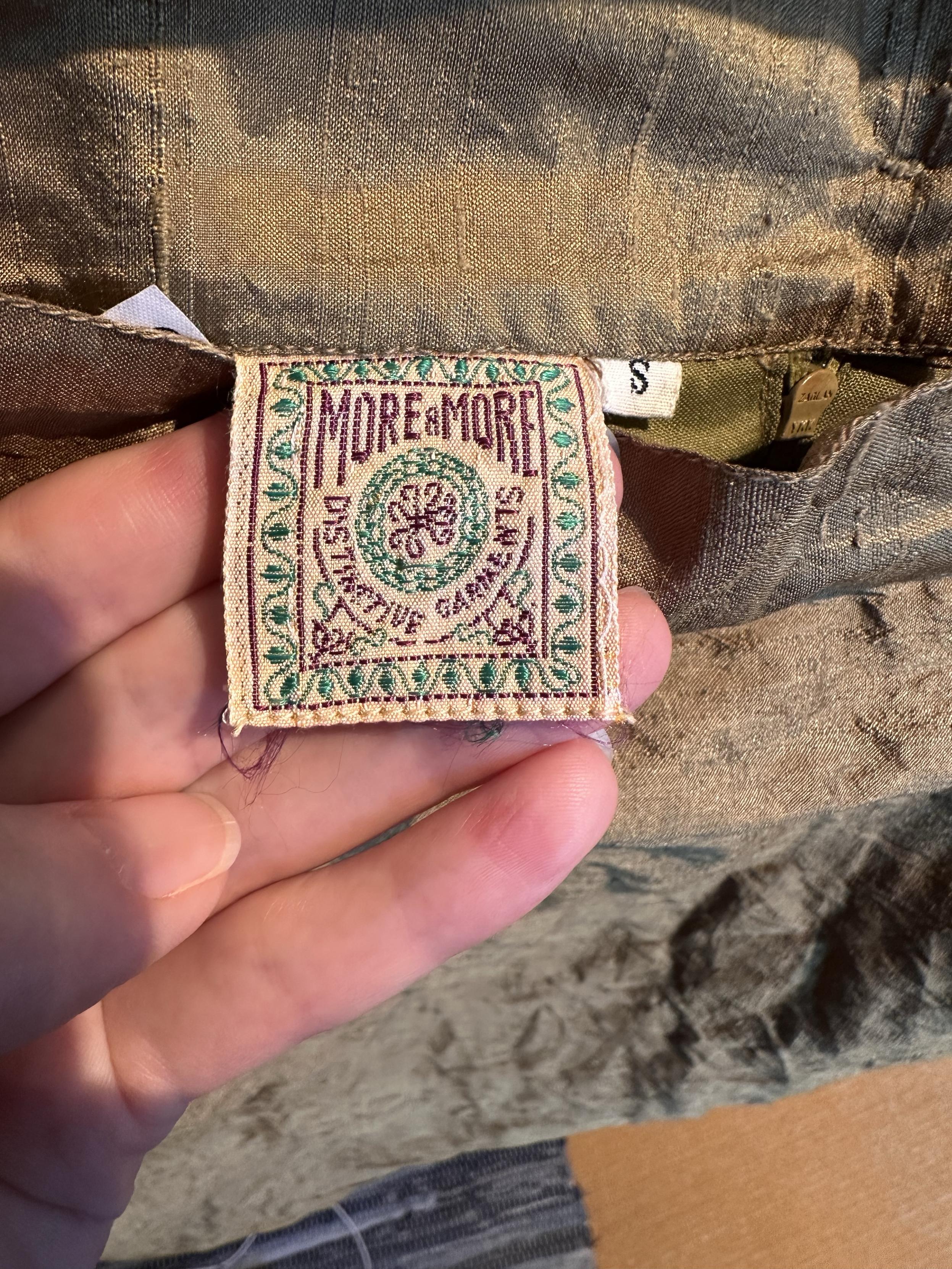 Silk skirt’s label. It’s machine embroidered in Christmas tree green and Bordeaux. It says “More & More distinctive garments”, written around a circular design, and framed by a square border decorated with vines.