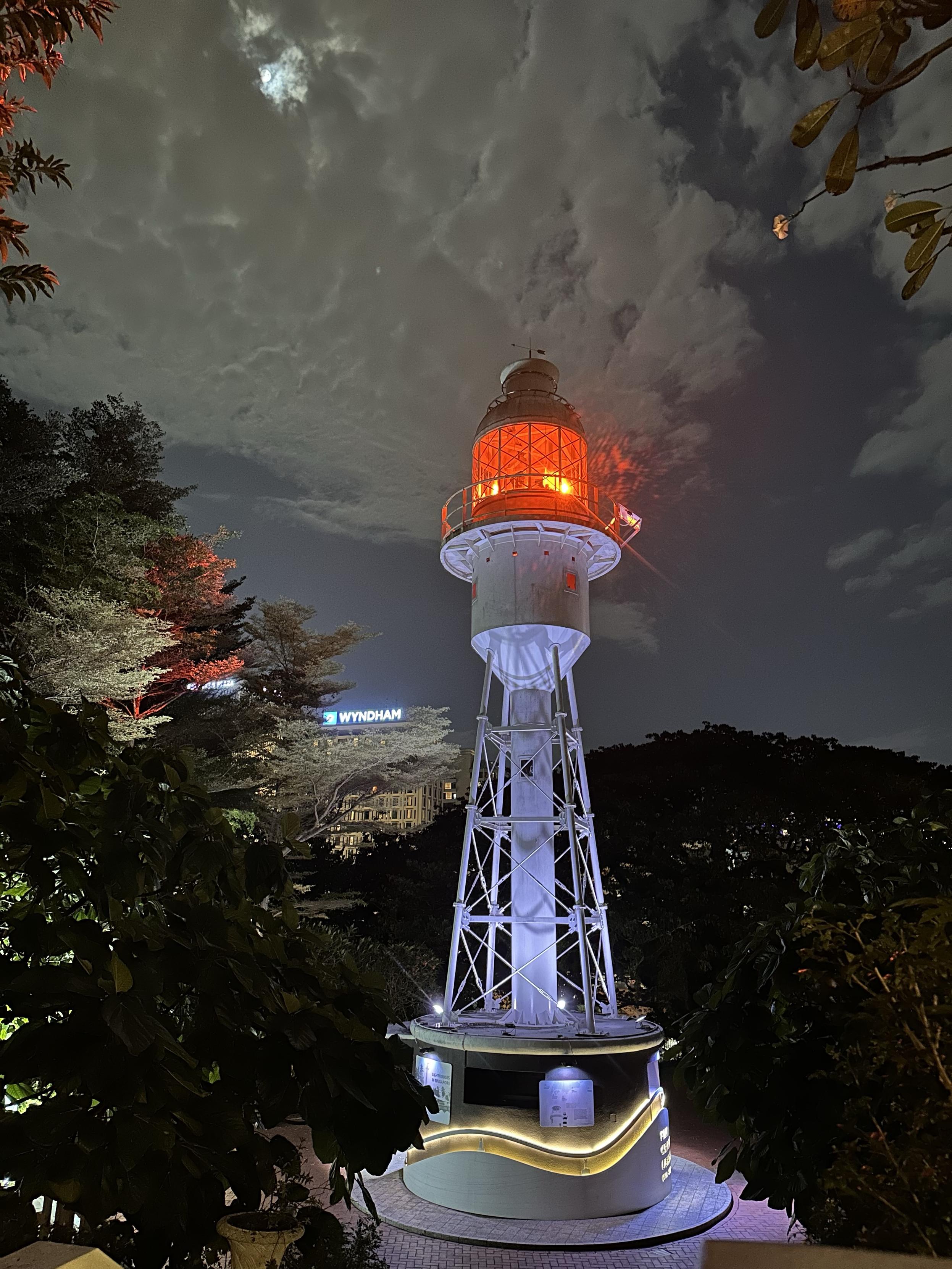 Fort Canning lighthouse, seen from sight higher up the hill. Is an open structure of white metal, with a red light in the top. In the background are trees and a few tall buildings. The sky is mostly overcast, but the full moon is peeking through the clouds.