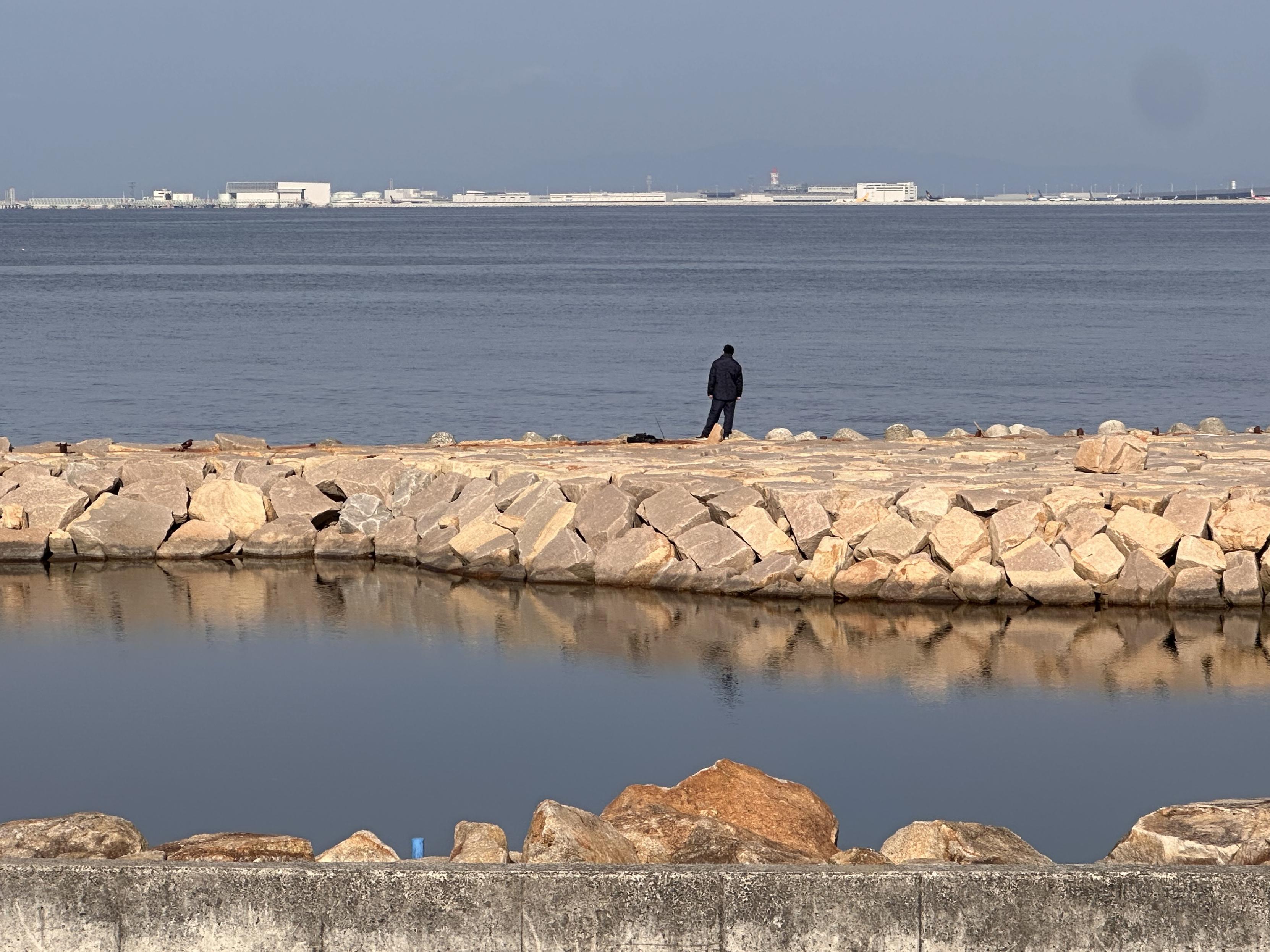 A lone man stands on a man-made stone barrier, looking out over the water to the airport in the other side of the bay. Blue sky, sunny