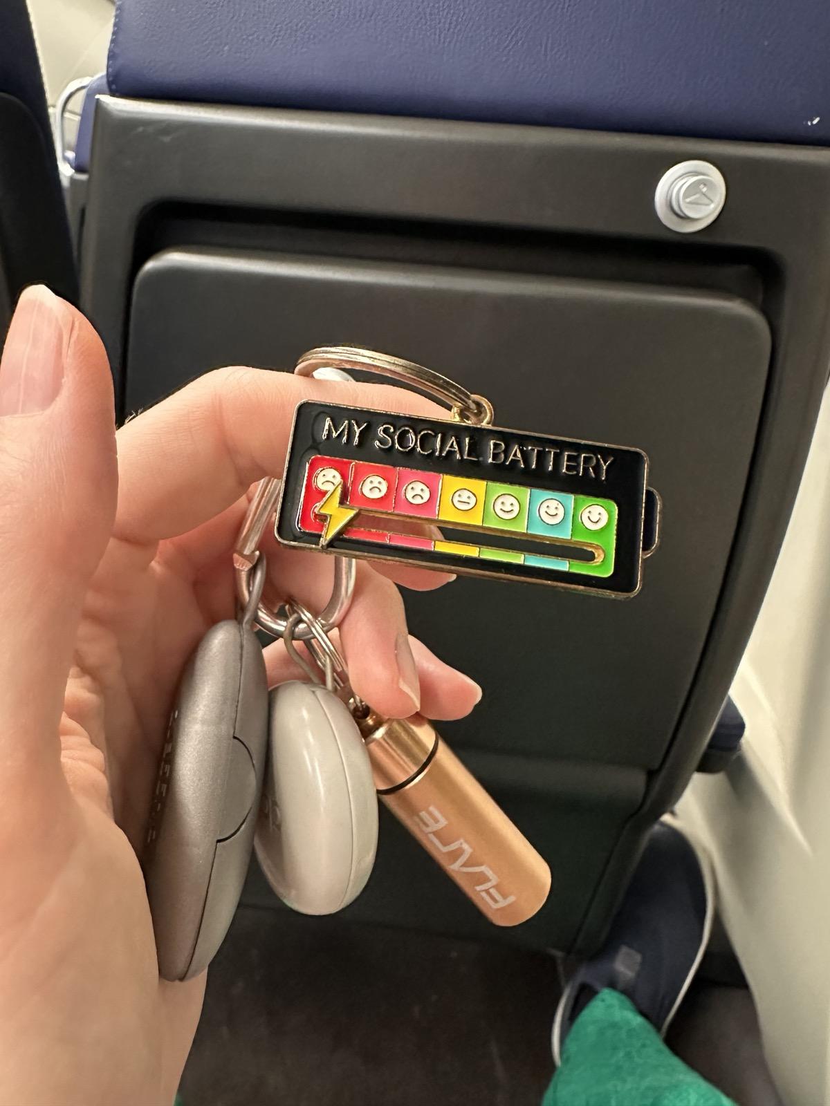 My hand holding a key chain that says "My Social Battery", and the lightning-shaped slider is all the way to the left, in the red with an angry face. Key chain attached to a bundle of ear plug cases (pluggerz, loop, flare).