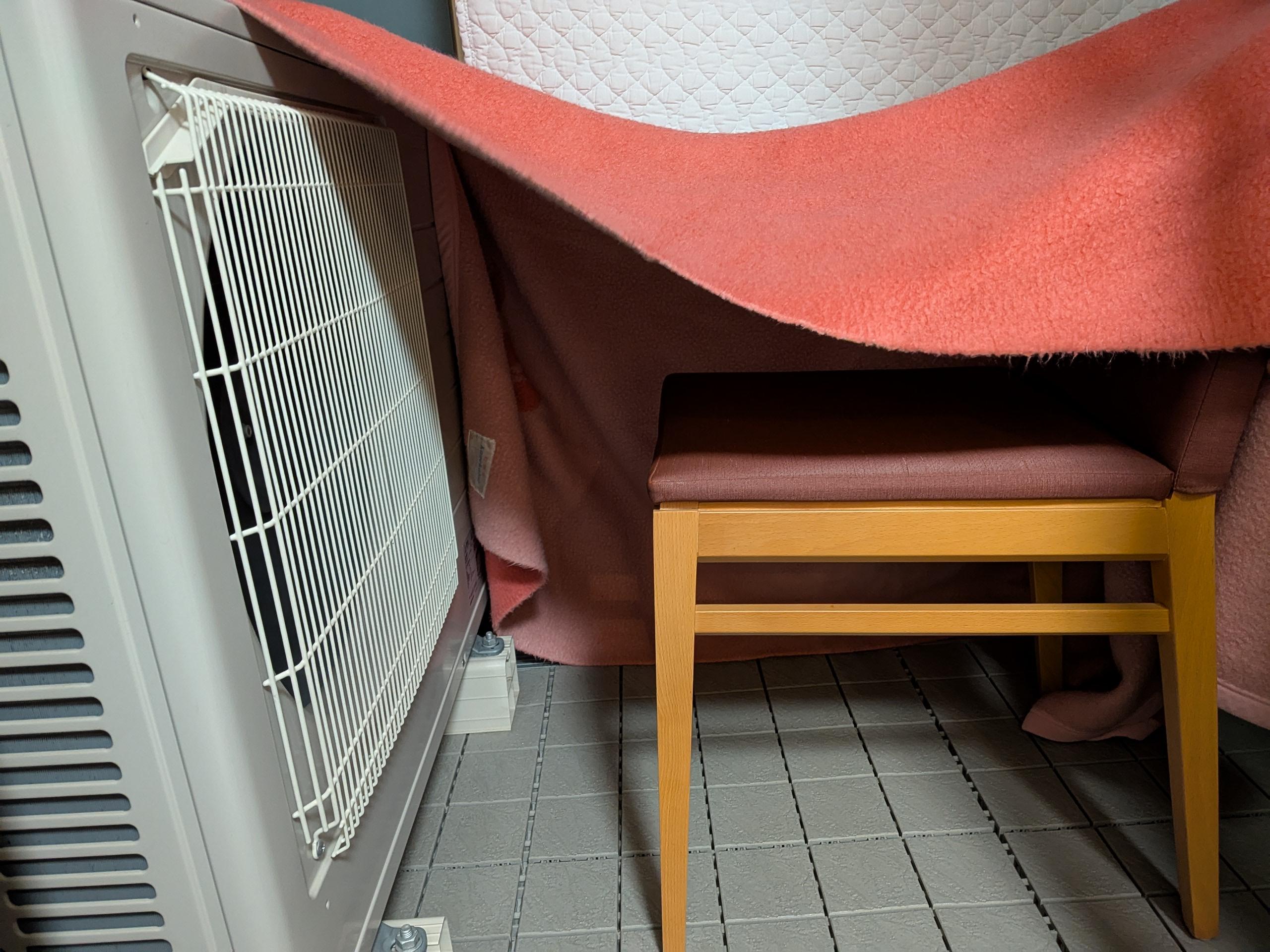 A chair faces the fan unit of a heat pump. A blanket covers 3 sides, leaving only the side facing the camera open.