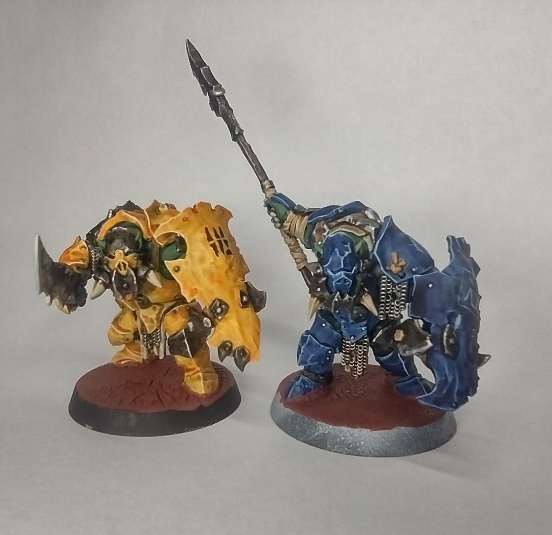 Painted miniatures of two orcs, one on the left with a bright yellow armour, and one on the right with a dark blue one.