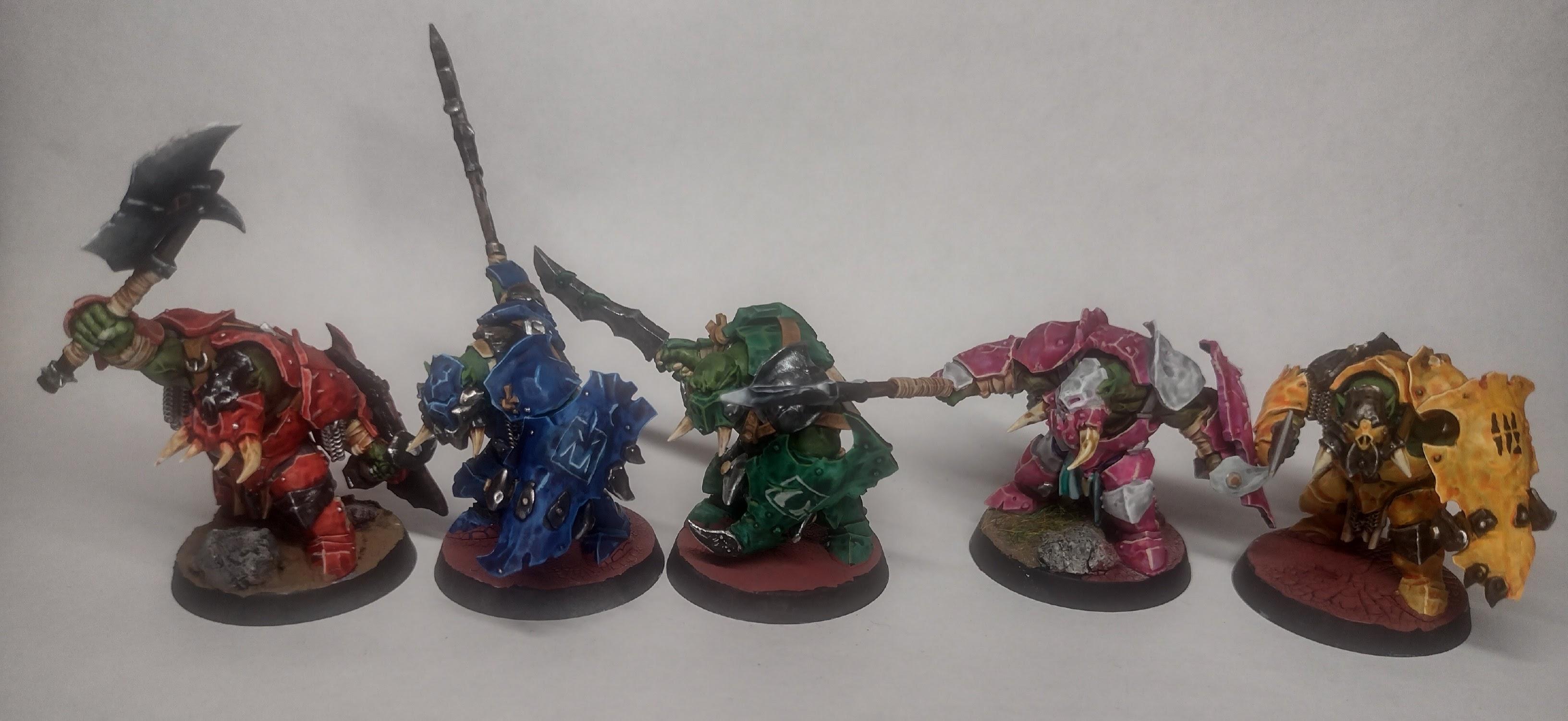 5 painted orc miniatures, one in red armour, one in blue, one in green, one in pink, and one in yellow. Placed in a line.