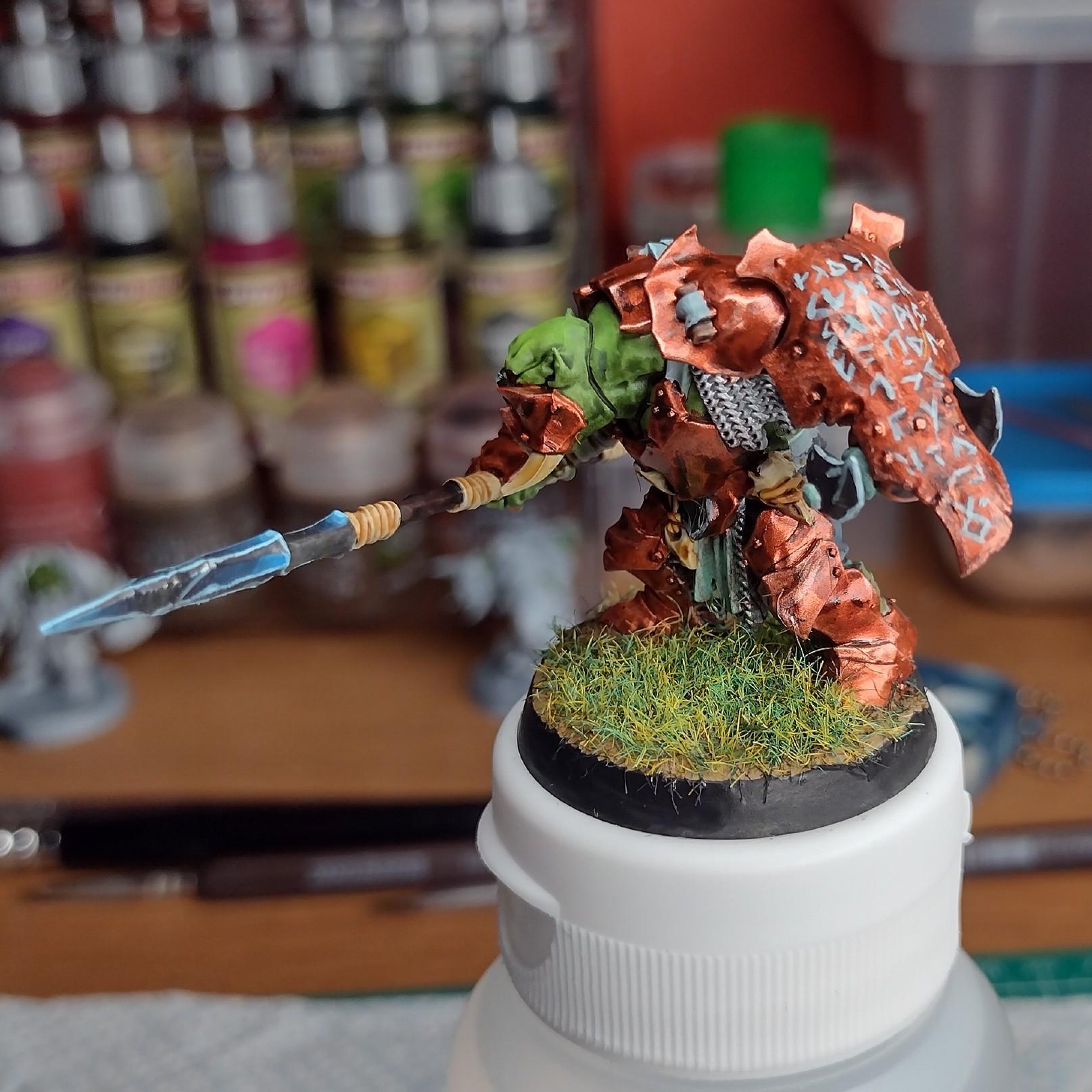 Orc miniature painted with a copper armor. In one hands it holds a big spear, and in the other a large shield covered in runes.