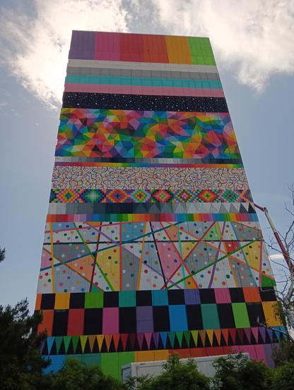 Mural "In Equality" of the Spanish artist Okuda San Miguel on the APA tower in Vienna
The painting has vibrant colors, and different patterns. Description of rows from top to bottom:
Rectangles (violet, pink, red, orange, yellow, light green)
Horizontal stripes, white, grey, pastel blue, pastel pink, black with with dots reminiscent of stars
A large section with a mosaic with lots of small triangles with different colors
Small stripe with greyscale
Small stripes with rainbow spectrum
A section …