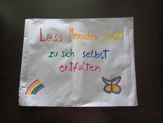 Zwei zusammengeklebte, weiße A4-Seiten bemalt mit farbigen Filzstiften mit dem Text "Lass Menschen sich zu sich selbst entfalten". Im unteren linken Eck gibt es einen kleinen Regenbogen mit rot, orange, gelb, grün, blau, violett. Der Text ist auch mit diesen Farben geschrieben. Im unteren rechten Eck ist ein Schmetterling gemalt, mit schwarzem Umriss, braunen Fühlern. Die oberen Flügel sind pink, weiß, hellblau. Die unteren Flügel sind am Rand pink und hellblau und großteils gelb mit einem klei…