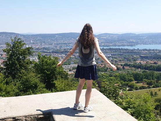 A photograph of me from behind. I'm standing on the edge of the wall of the ruins of the Friesenberg castle. Below the wall are some trees and bushes and farther away Zurich and Lake Zurich are visible. I'm wearing white sneakers, a short blue skirt, a gray shirt. My hands are stretched the the side slightly backwards, my long chestnut brown curly hair is nicely falling along my upper back.