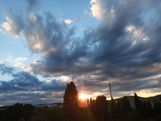 Photo of a sunset at the northern end of Vienna. There are swathes of clouds, in the process of loosening up, partly illuminated, partly shaded dark blue with light blue sky visible on the left and top center. The sun is glowing orange in the lower center between some trees, with some rays and a red oval lens flare below. Between the trees are some buildings with hills behind them.