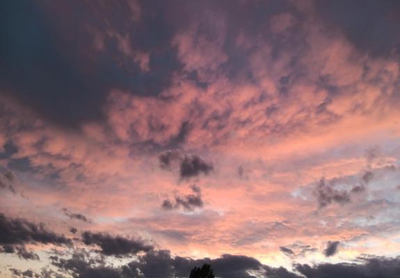 Photo of clouds illuminated by the setting sun. The topmost layer of clouds is rather bumpy and has a pinkish-orange color, with shades intermixed and the upper left and right corners are completely shaded. In the middle, the clouds are a bit more orange, and bits of light blue sky show behind some holes there. A few fully shaded clouds are in front of the middle and the bottom of the image, the ones in the middle small and dispersing. On the bottom, right and center, close to the sun, there ar…