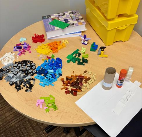 An office desk, with piles of Legos sorted into piles by color; some paper, cardboard tube, glue stick, and tape; the textbook "Artificial Intelligence: a Modern Approach"