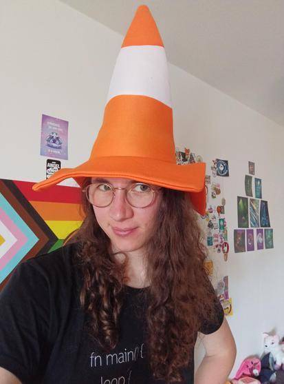 Selfie of me wearing an orange-white VLC street cone as a hat. I'm wearing a black shirt, round glasses. I have grey-green eyes, a satisfied look and a slight smile. My long chestnut brown curly hair is slightly reddish, because of the sun/lighting. In the background are some posters on the wall and plushies on the couch.