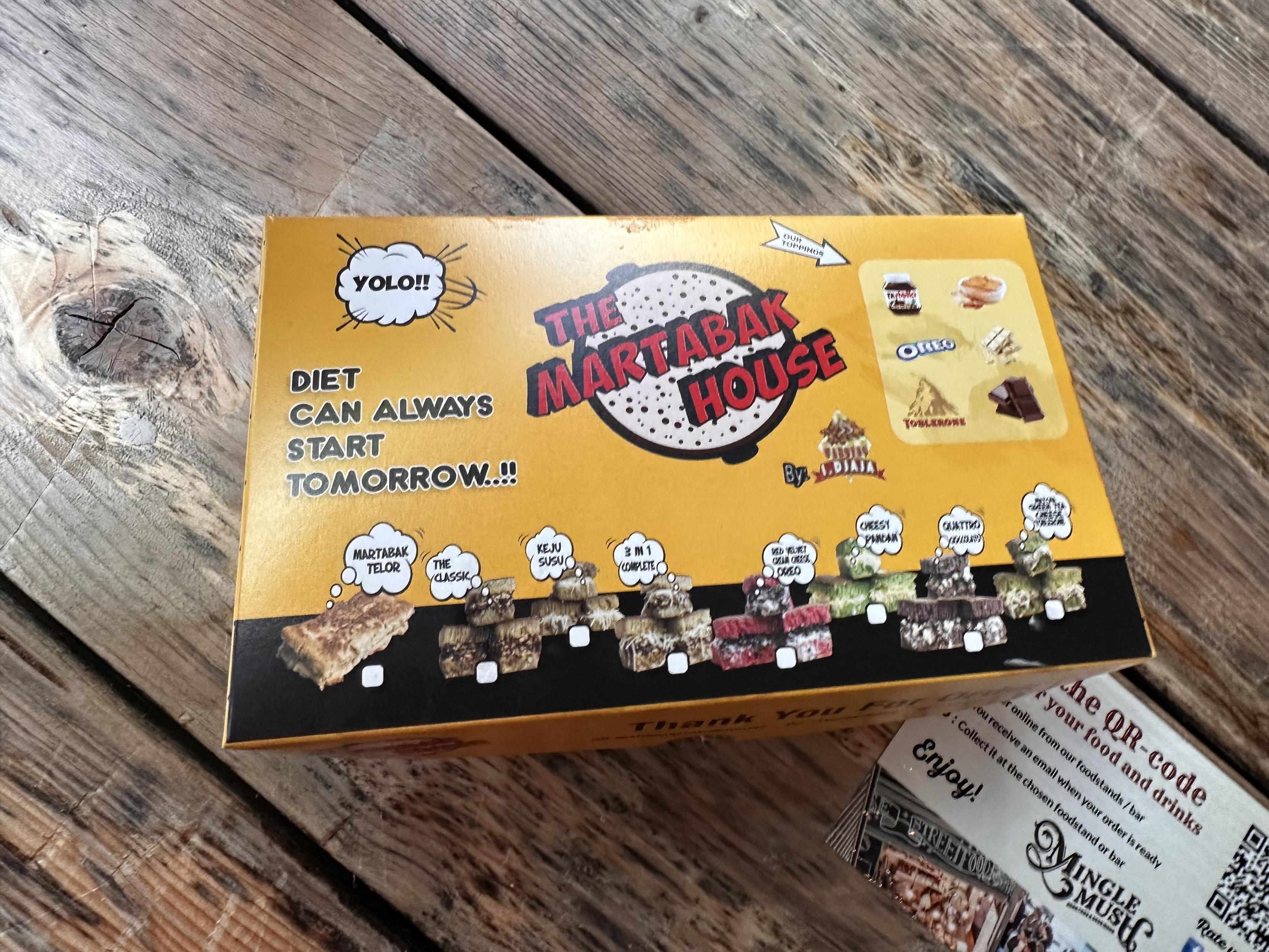 A bright yellow cardboard takeaway box from "The Martabak House" on a wooden table. The box says “YOLO - diet can always start tomorrow”.  it features pics of various toppings and various martabak options.