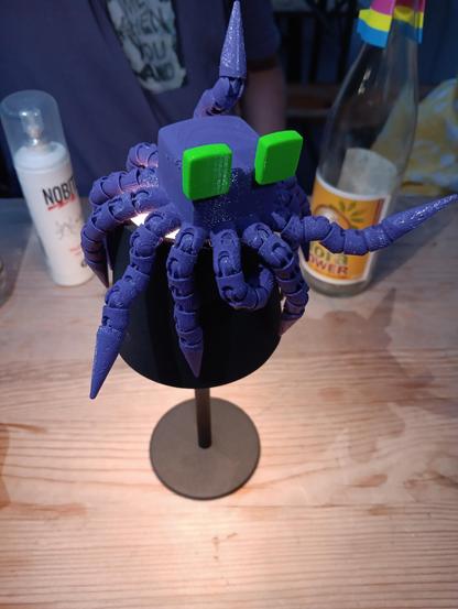 Blob-Maskottchen vom Håck Ma's Castle (3D-gedruckte Figur mit vielen biegbaren Tentakeln aus violettem Filament und 2 quadratischen, grünen Augen), was auf einer schwarzen, kleinen Tischlampe steht. Das Filament schimmert ein bisschen im Licht was von oben drauf leuchtet, die Lampe leuchtet unterhalb.