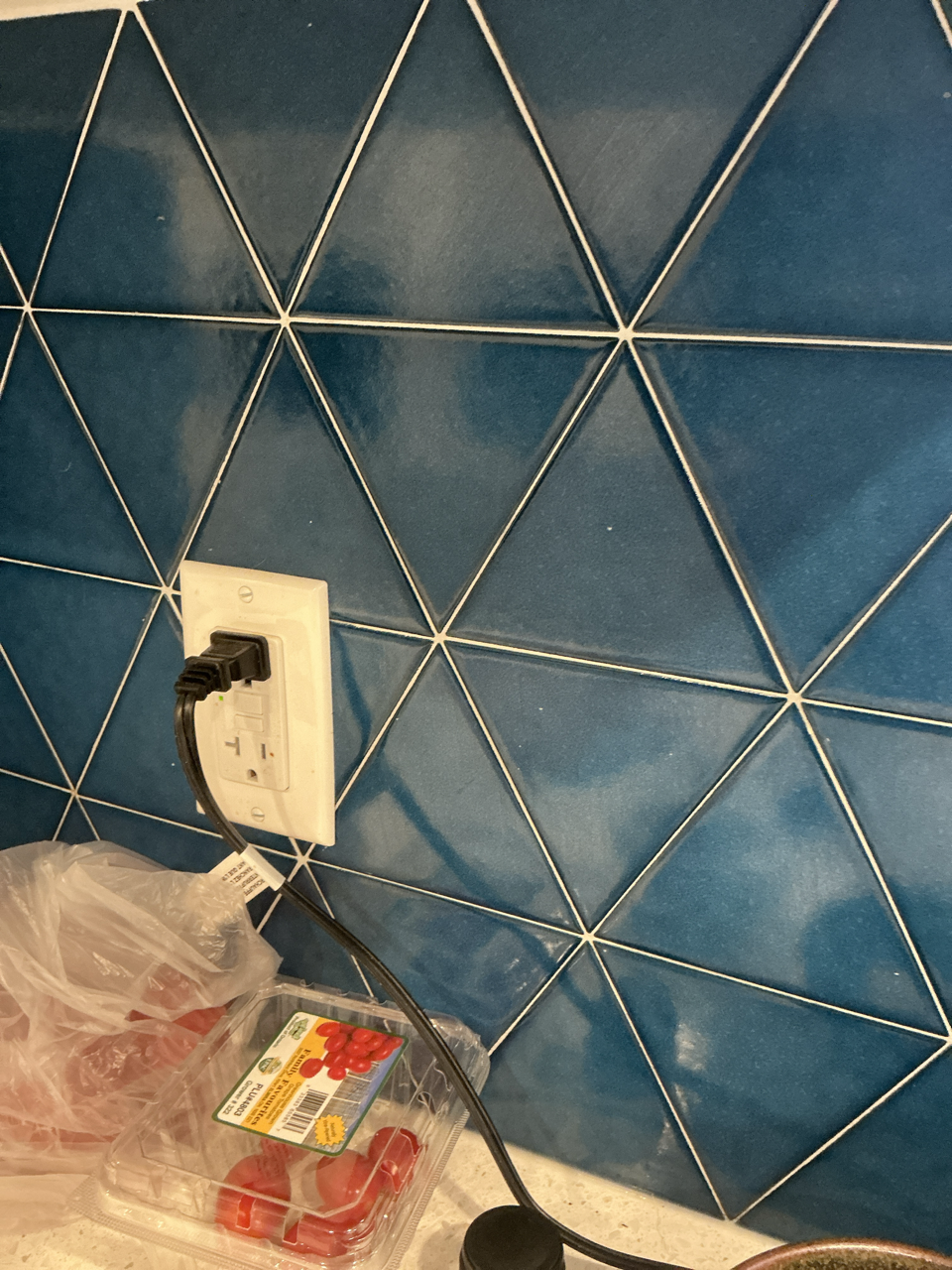 A photograph of a kitchen backsplash, showing royal blue glazed ceramic tiles in the shapes of equilateral triangles. The backsplash is interrupted by a North American electrical outlet, and various tomatoes are visible on the counter below.