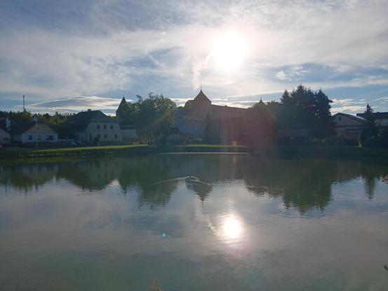 Der untere Teich in Ottenschlag, Richtung Morgensonne, die über dem breiten Südwestturm vom Schloss Ottenschlag aufgeht, der zusammen mit einem Stück Südmauer in der hinteren Mitte des Bildes zu sehen ist. Daneben sind andere Häuser und ein paar Bäume zu sehen. Im Wasser ist die Reflektion davon zu sehen und von der Sonne und den lichten Wolken, die sie umgeben. In der Mitte vom See schwimmt eine Ente, Richtung Schloss und hinterlässt seitlich kleine Wellen.