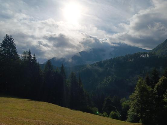 Dark green trees on hills and mountains with the sun shining from behind some clouds, that are covering part of the mountain and parts of the sky. In front is a dry meadow, going downhill.