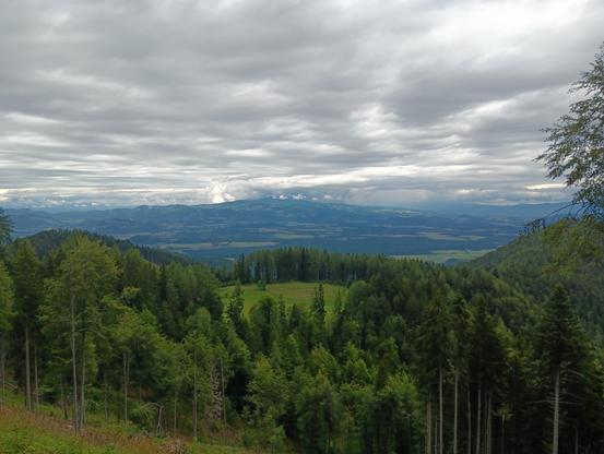 View towards the north of Carinthia. The sky is full of clouds, with some bright white parts in between greyer areas. In front are hills covered by trees and a meadow on the middle hill. Further back is lower with some villages and forests. In the very back there's mountains again, in a bluish tint.