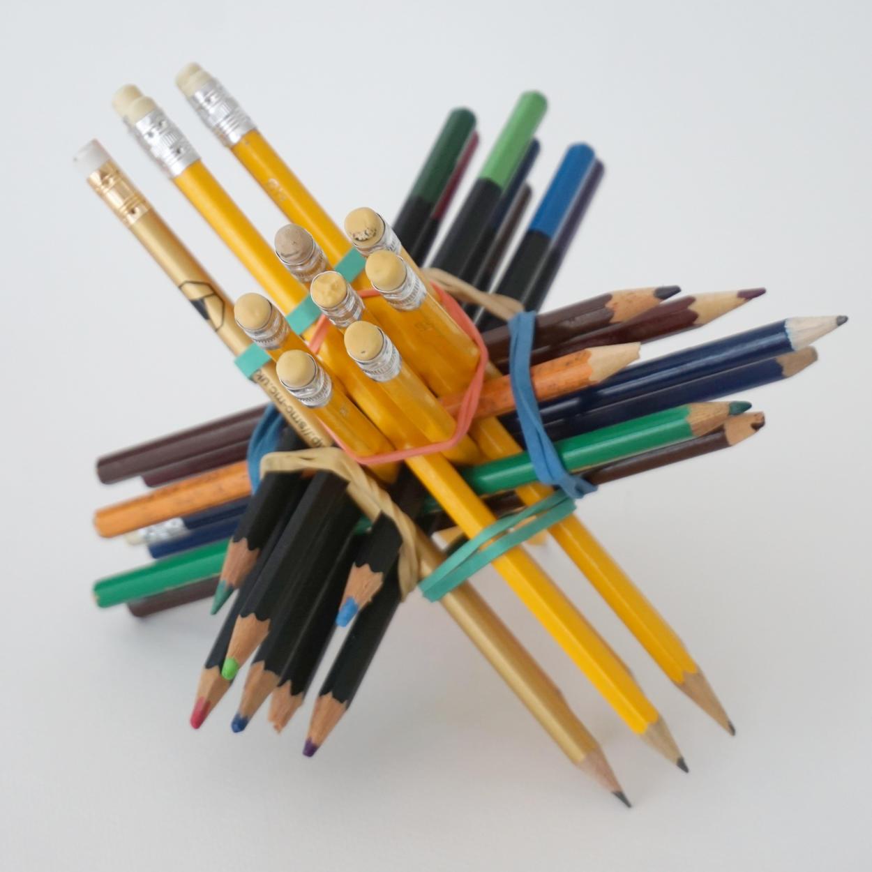A structure made of pencils arranged in four sets of seven pencils that are held together with rubber bands. Each set of pencils points in a different direction and are mostly similar, e.g. rubber-tipped yellow pencils and colour pencils of different kinds.