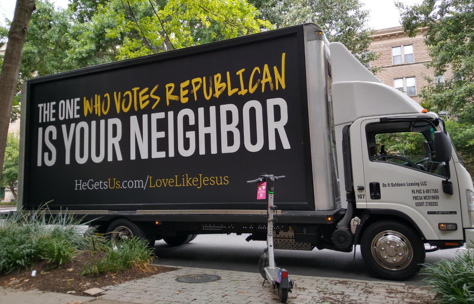 A large truck has a sign on its side that reads

THE ONE WHO VOTES REPUBLICAN
IS YOUR NEIGHBOR 

HeGetsUs.com/LoveLikeJesus

The background is grey/brown.

The text is white except Us and LoveLikeJesus are yellow text and WHO VOTES REPUBLICAN uses a yellow hand painted writing style

The truck is surrounded by green leafy trees and an electric scooter is on the sidewalk pointing towards the truck.